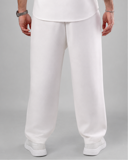 A person shown from the back wearing loose white trousers, a white top, and white sneakers against a light gray studio background.