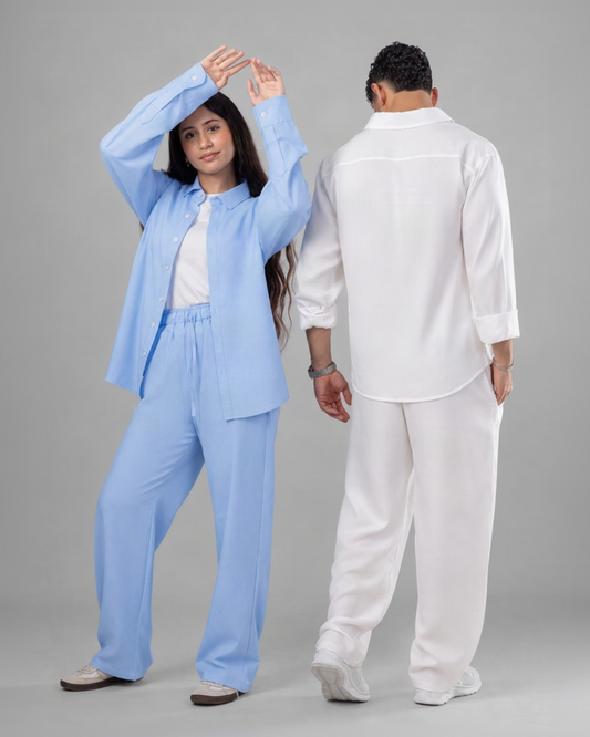 A man and a woman modeling matching lounge sets against a solid gray studio background. The woman on the left wears a light blue button-up shirt and matching wide-leg pants over a white top, while the man on the right wears a white button-up shirt and matching white pants.