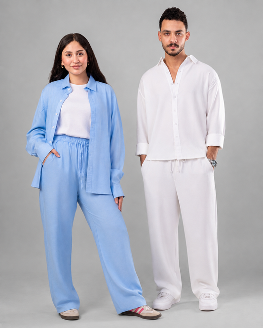 A man and a woman modeling matching lounge sets against a solid gray studio background. The woman wears a light blue button-up shirt and matching wide-leg pants over a white top, while the man wears a white button-up shirt and white wide-leg pants.
