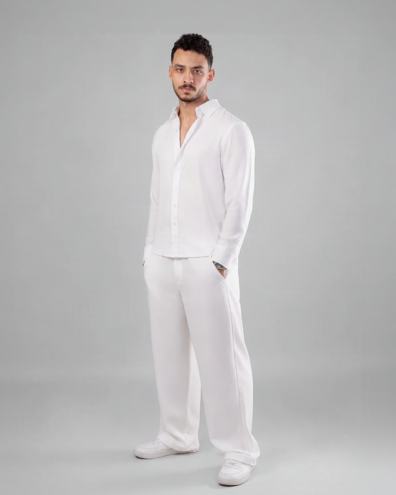 A man modeling a white long-sleeve button-up shirt and matching white trousers against a solid gray studio background.