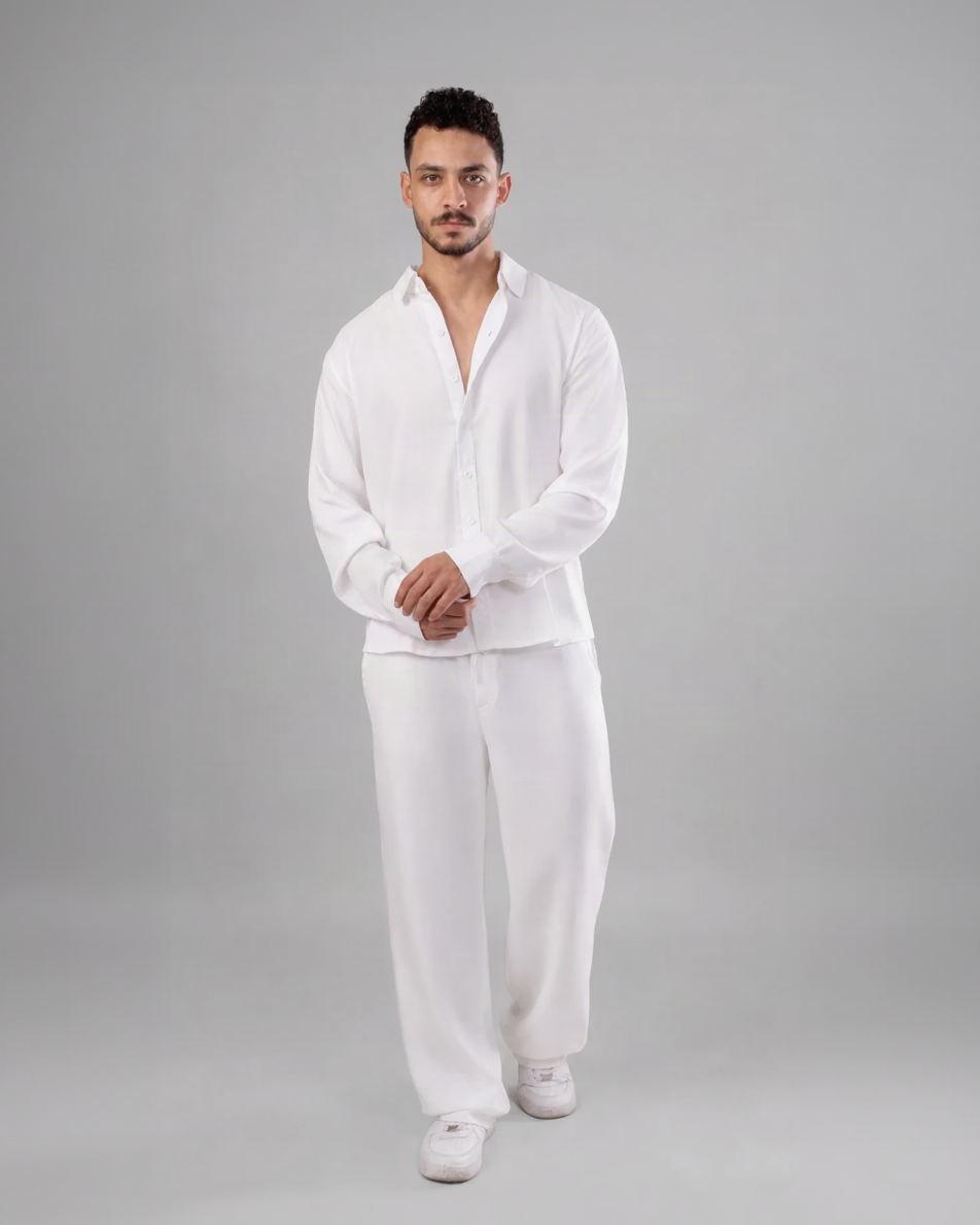 A man modeling a white two-piece lounge set consisting of a long-sleeve button-up shirt and matching wide-leg pants, paired with white sneakers, against a solid gray studio background.