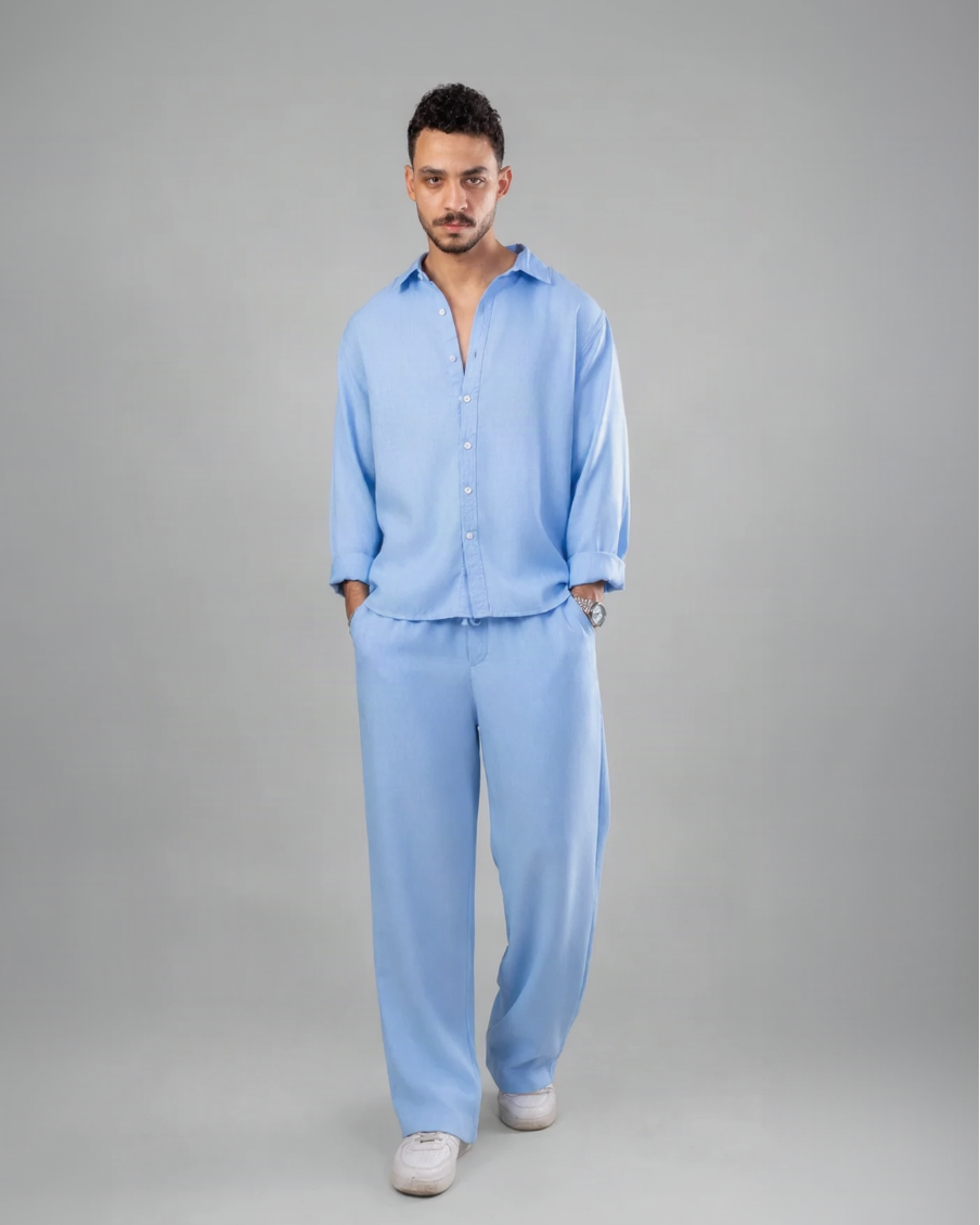 A man modeling a light blue two-piece lounge set consisting of a long-sleeve button-up shirt and matching wide-leg trousers, standing against a neutral gray studio background.