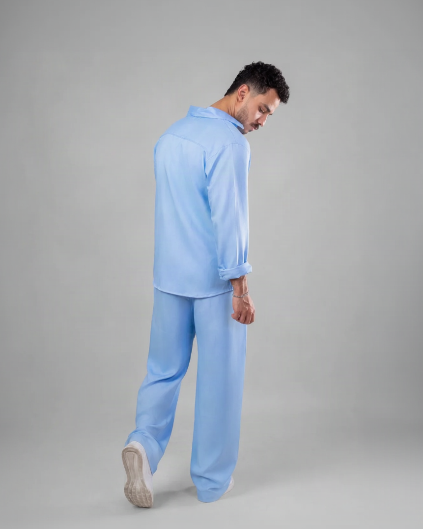A man wearing a light blue long-sleeve shirt and matching wide-leg trousers, shown from a side-back angle against a plain gray studio background.