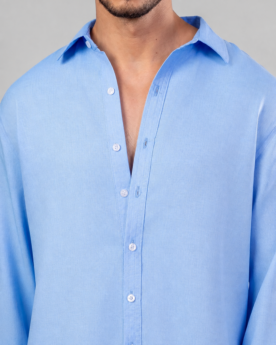 A close-up of a man wearing a light blue long-sleeve button-down shirt, showing the collar, front buttons, and chest area against a neutral gray background.