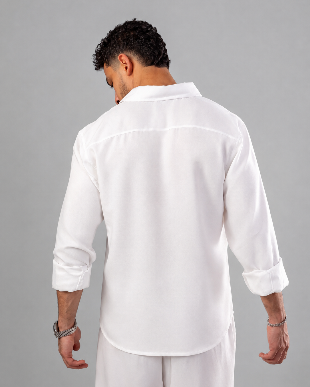 A man viewed from behind wearing a white long-sleeve button-up shirt and matching white trousers against a solid gray studio background.