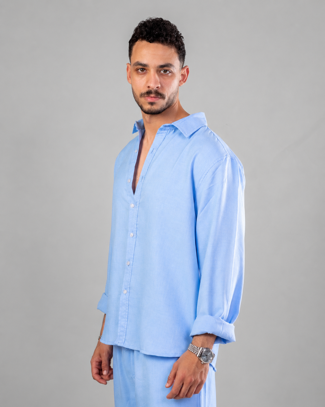A man modeling a light blue long-sleeve button-up shirt and matching light blue pants against a solid gray studio background.