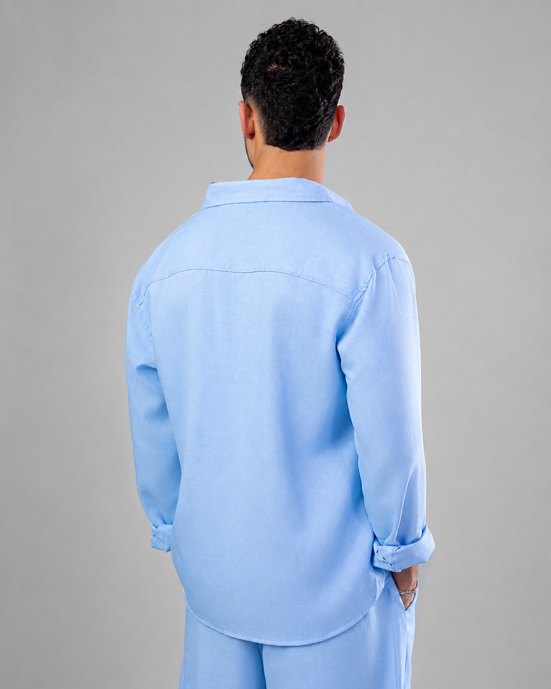 A man viewed from behind wearing a light blue long-sleeve button-up shirt and matching light blue pants against a plain gray studio background.