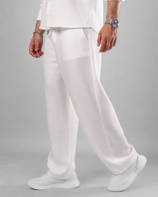 A person wearing loose white trousers with a matching white top and white sneakers, photographed against a light gray studio background.