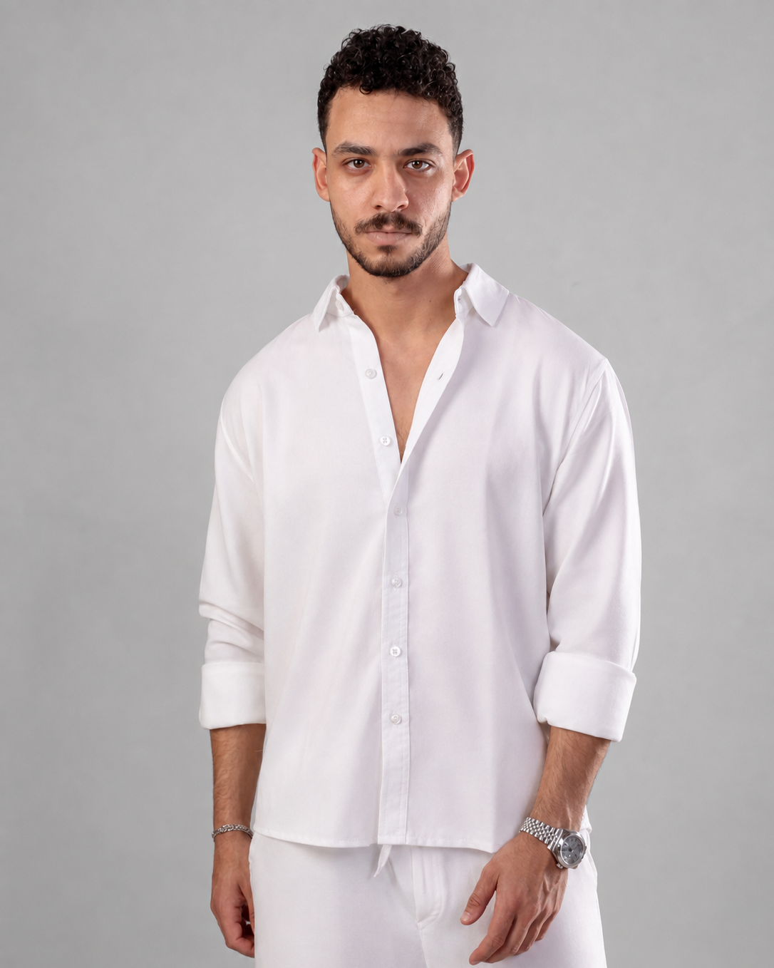 A man wearing a white long-sleeve button-down shirt with rolled-up cuffs and white trousers, standing against a solid gray studio background.